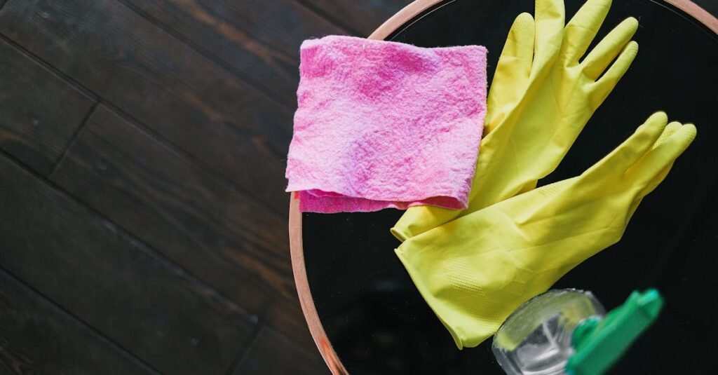 Yellow gloves and pink cloth on a table with cleaner bottle, viewed from above.