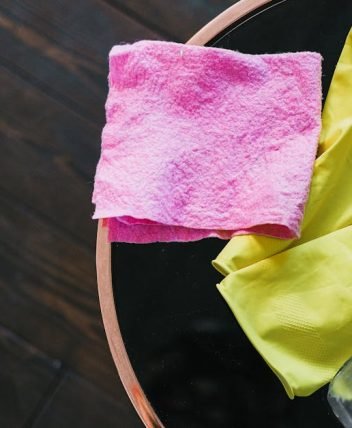 Yellow gloves and pink cloth on a table with cleaner bottle, viewed from above.